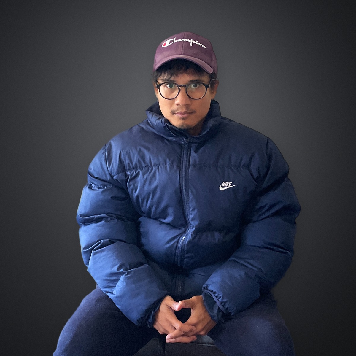 Travis Yangzte in a maroon Champion cap, glasses and a navy Nike puffer on a charcoal studio backdrop