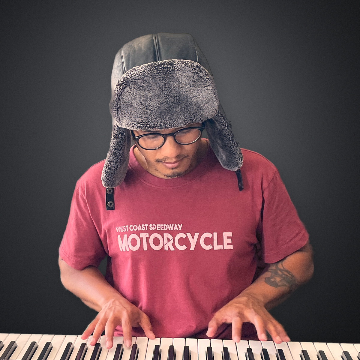 Travis Yangzte in a fur trapper hat, glasses and a red tee, playing a keyboard in the studio